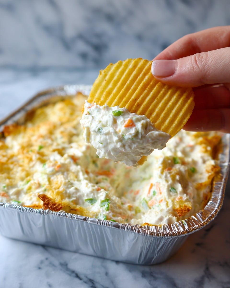 A close-up shot shows a woman's hand holding a yellow ridged potato chip dipped in a thick white creamy dip with visible small orange and green bits, suggesting a mix of ingredients inside the dip. The dip fills a silver foil tray, covering the whole base with a slightly uneven texture. The background is a white marbled surface, softly blurred to keep focus on the chip and dip. photo taken with an iphone --ar 4:5 --v 7
