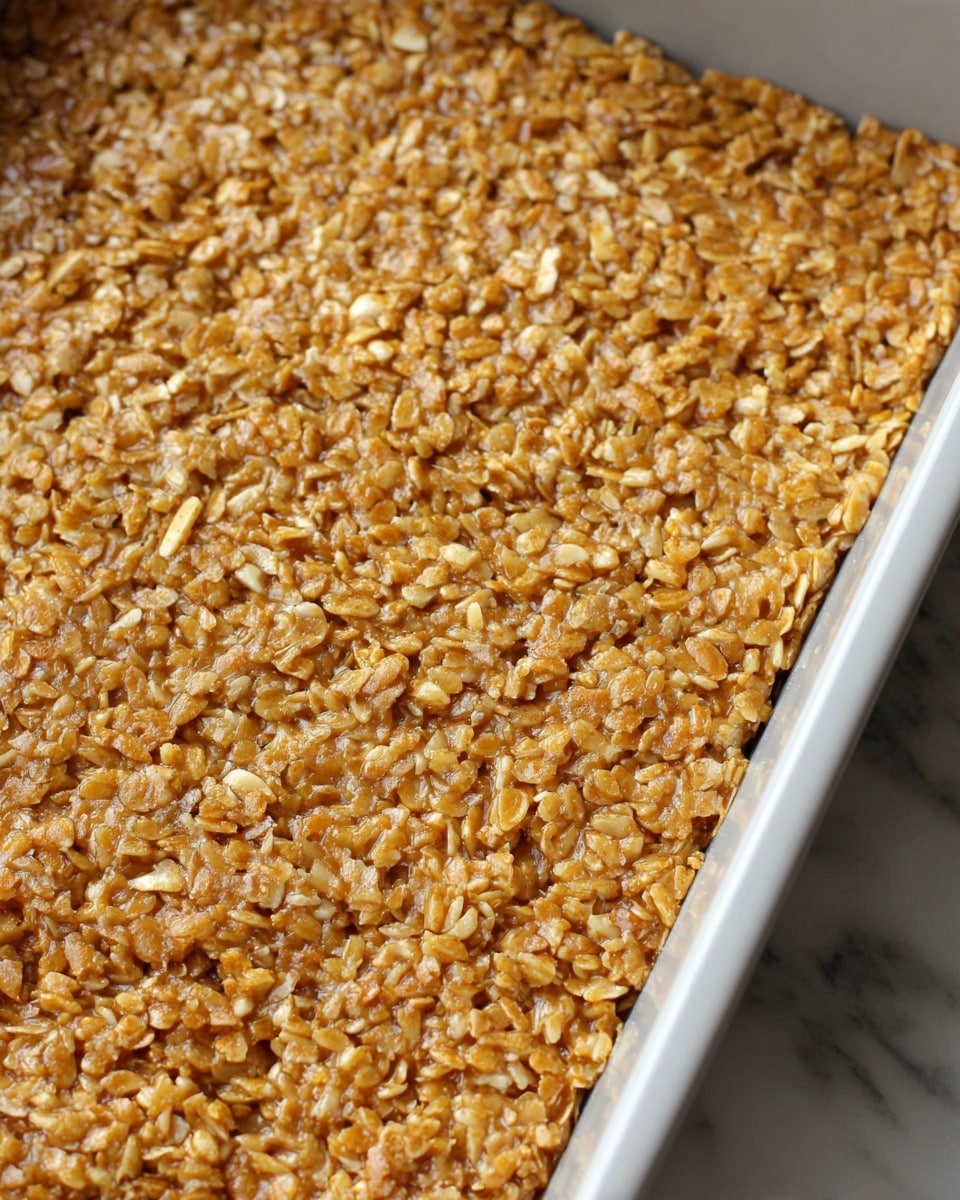 A close-up view of a thick, single-layered dessert bar in a white rectangular baking pan, showing a mix of light to medium golden brown rice and nut pieces tightly packed together, with a slightly shiny, sticky texture covering the surface; the pan is placed on a white marbled surface. photo taken with an iphone --ar 4:5 --v 7