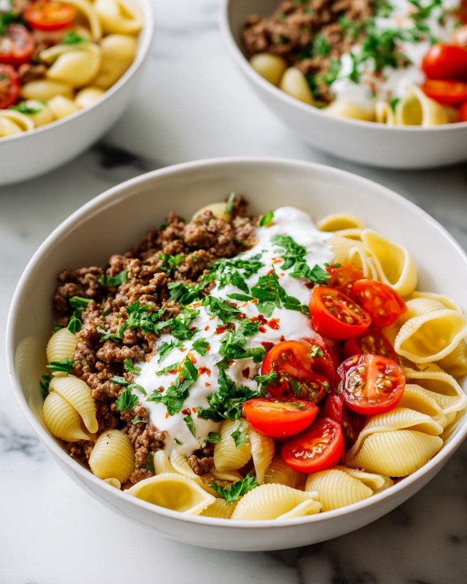 A white bowl filled with three layers: at the bottom, light yellow shell pasta with a smooth, curved texture; in the middle, a dollop of white yogurt sauce spread in a line; on top, browned ground meat mixed with finely chopped onions, bits of red chili oil, fresh green chopped parsley scattered throughout, and bright red cherry tomato halves placed mainly in the center. The bowl is set on a white marbled surface, and parts of two other white bowls with similar ingredients are visible at the top edge of the image. Photo taken with an iphone --ar 4:5 --v 7