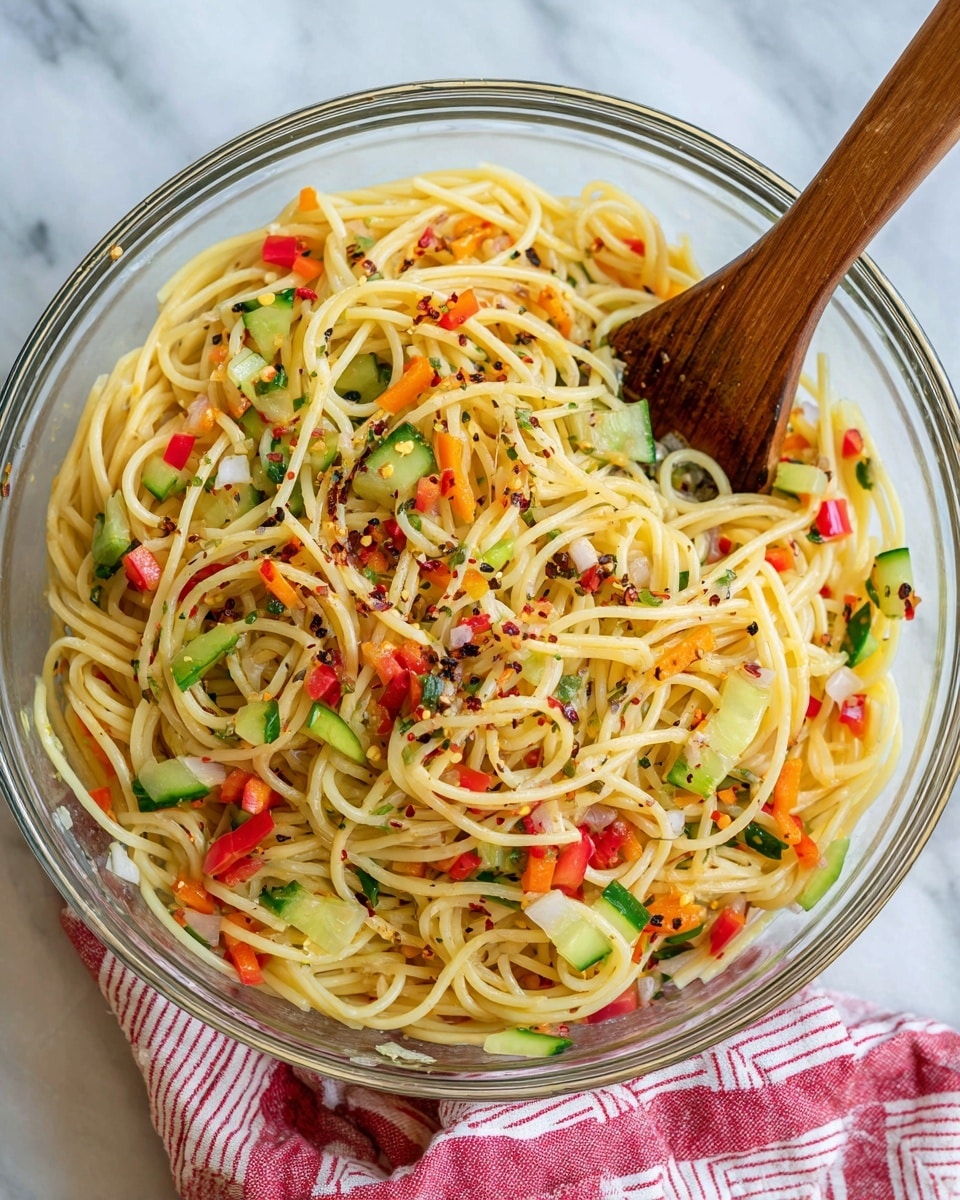 The image shows a clear glass bowl filled with a colorful spaghetti salad. The dish has one main layer of long, light yellow spaghetti noodles, mixed evenly with small pieces of green cucumber, red and orange bell peppers, and diced onions. The noodles and vegetables are sprinkled with black pepper and red chili flakes, giving the dish a speckled look. A wooden spoon pokes in from the left side, resting inside the bowl. The bowl is placed on a white marbled surface, with a red and white striped cloth partially visible underneath on the left side. Photo taken with an iphone --ar 4:5 --v 7