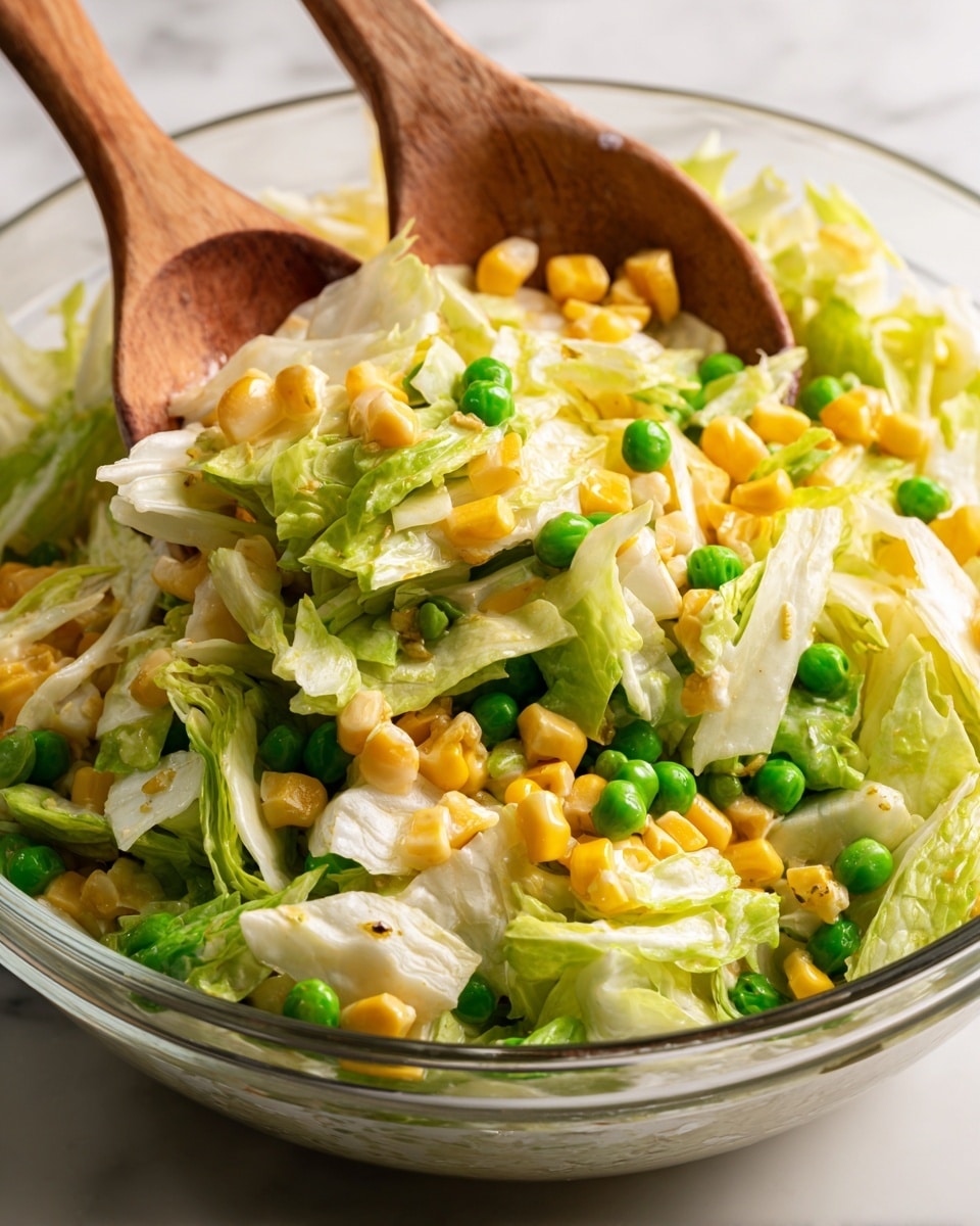 A close-up image shows a fresh salad being lifted with two wooden spoons. The salad has three main layers: pale green leafy napa cabbage with a crisp texture, bright yellow corn kernels with smooth round shapes, and vibrant green peas scattered evenly. The salad looks creamy with a light dressing coating the vegetables, giving a slight shine. The background is a white marbled surface, and the salad sits in a clear bowl. photo taken with an iphone --ar 4:5 --v 7