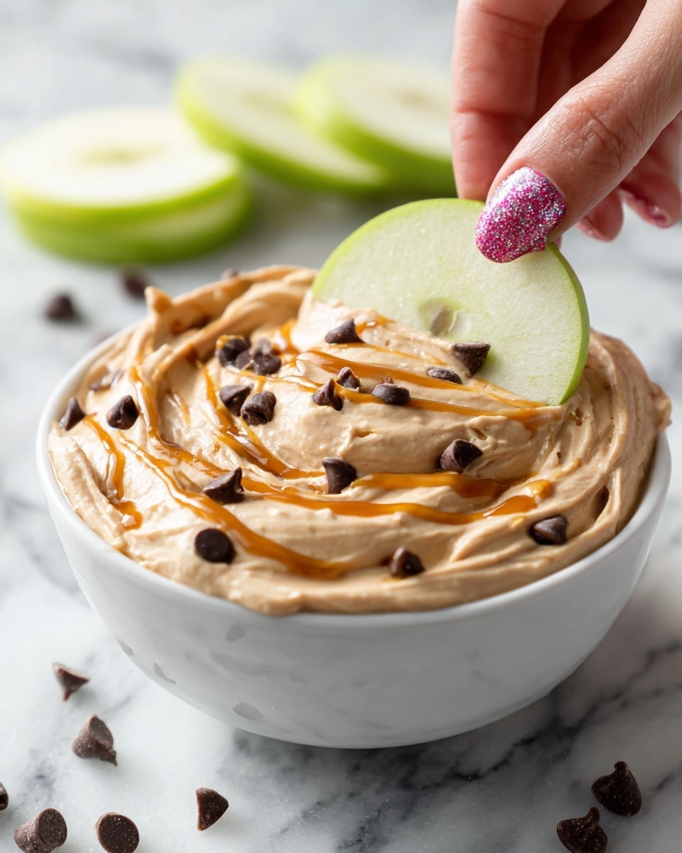 A white bowl filled with creamy dip that is a light tan color with a smooth, thick texture. Dark chocolate chips are mixed into the dip and slightly visible on the surface. A drizzle of golden caramel sauce is spread on top in thin, uneven lines. A woman's hand with glittery pink nail polish holds a bright green apple slice dipped into the creamy mixture. The bowl sits on a white marbled surface with some more green apple slices out of focus in the background and a few chocolate chips scattered nearby. Photo taken with an iphone --ar 4:5 --v 7