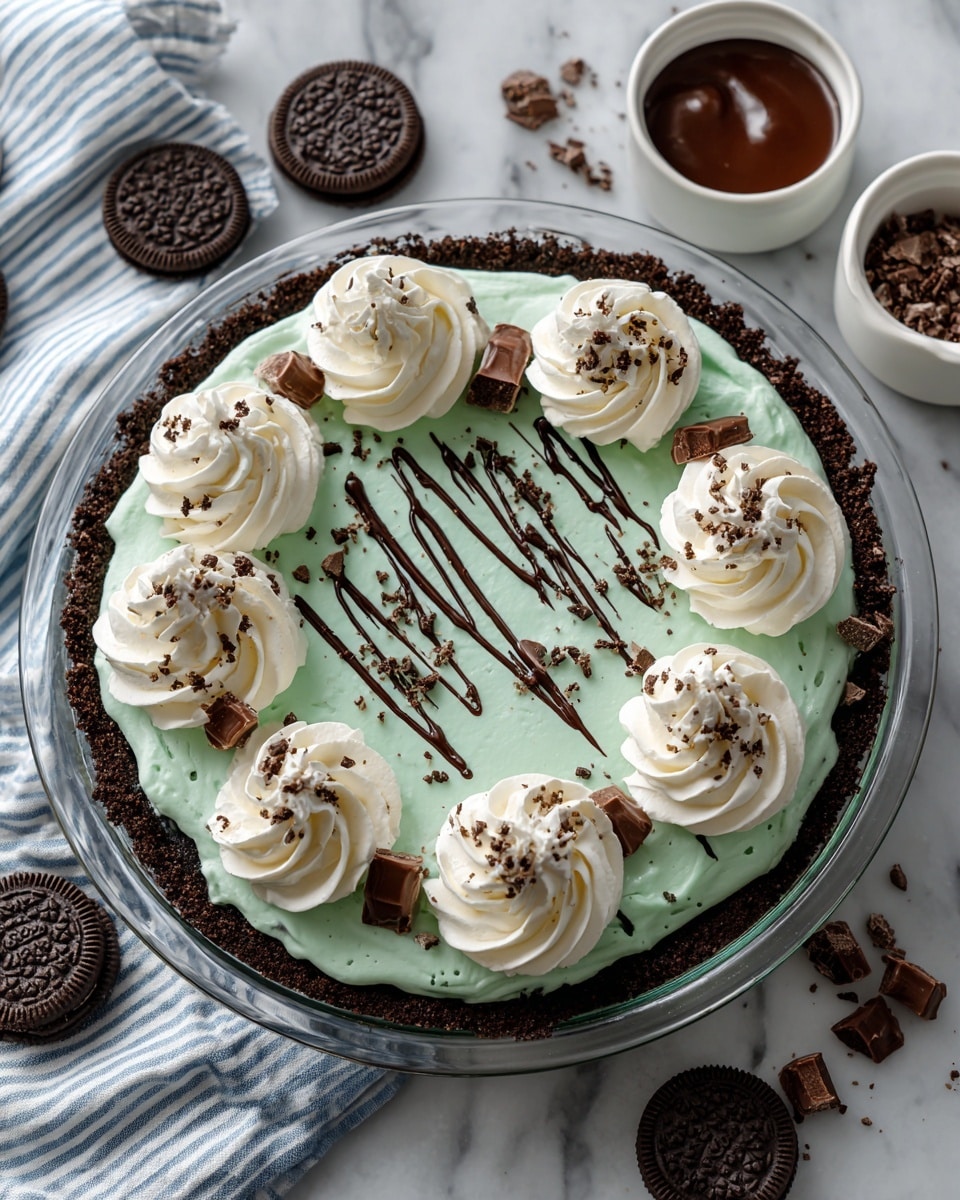 A dessert pie with three main layers is shown on a white marbled surface. The bottom layer is a dark chocolate crust pressed around the edges in a clear glass round pie dish. The middle layer is a smooth, light mint green filling spread evenly inside the crust. The top layer has white whipped cream swirls piped in a circle near the edge and one in the center. Each swirl is decorated with small pieces of chocolate candy. The middle green filling is drizzled with thin lines of dark chocolate and sprinkled with tiny chopped chocolate bits. Nearby on the surface are two Oreo cookies, crumbled cookie pieces, a striped white and blue cloth, and a small white bowl of chocolate sauce. Photo taken with an iphone --ar 4:5 --v 7