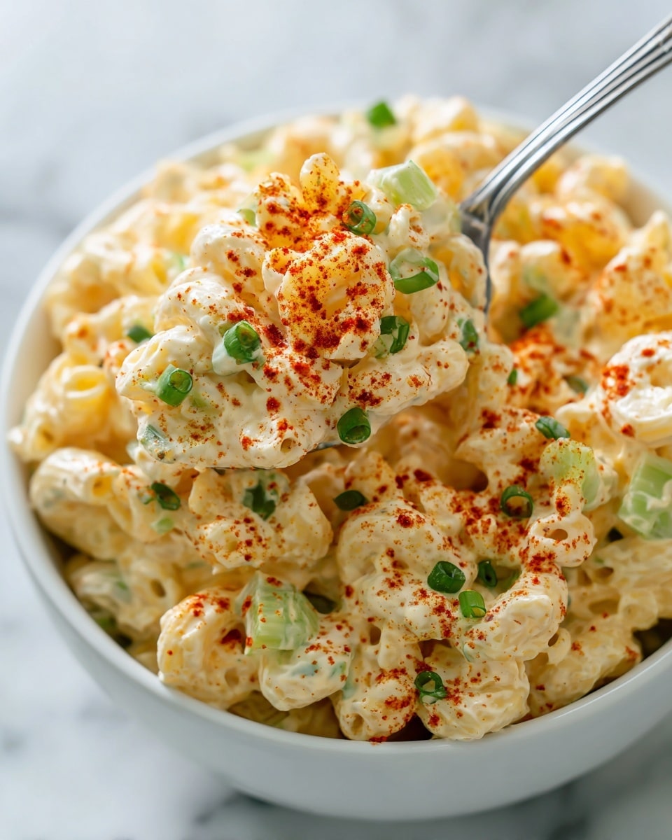 A close-up view of a white bowl filled with creamy macaroni salad, showing about two thick layers: the bottom layer is pale yellow pasta coated in a smooth, creamy dressing, and the top layer is sprinkled with bright red paprika and small pieces of chopped green onions that add green dots for color contrast; a silver spoon lifts a generous scoop of the macaroni salad, displaying soft pasta curls and bits of light green celery mixed into the creamy texture, all set on a white marbled surface. photo taken with an iphone --ar 4:5 --v 7