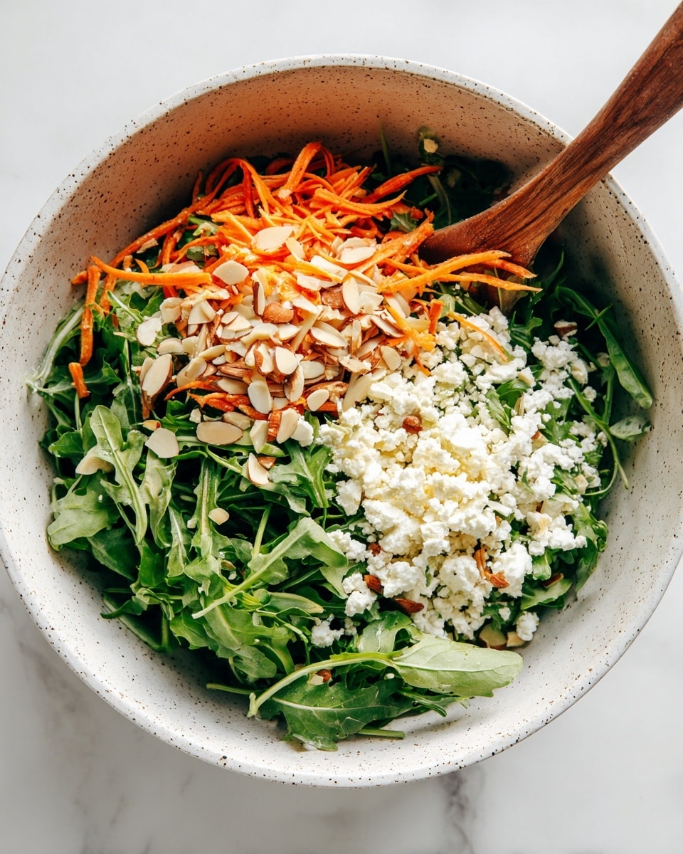 A white speckled bowl filled with a fresh salad is shown on a white marbled surface. The salad has three main layers: the bottom layer is green arugula leaves with a crisp texture, the middle layer consists of thin, curly orange carrot strips, and the top layer is scattered white crumbled cheese and light beige almond slices. A wooden spoon is partially visible inside the bowl, resting on the left side, mixing the ingredients. The colors are bright and natural, creating a fresh and healthy look. photo taken with an iphone --ar 4:5 --v 7