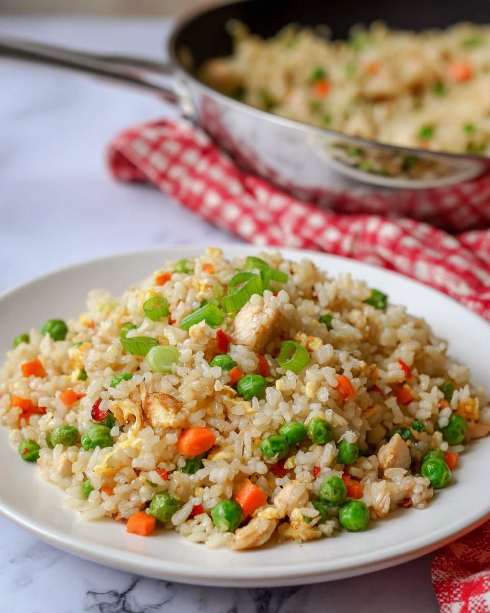 A close-up view of a white plate filled with fried rice showing three main layers: the base layer is light brown rice mixed with small pieces of cooked chicken; scattered throughout are bright green sliced green onions and diced green peas; sprinkled in small parts are red and orange bits of cooked vegetables. The texture of the rice looks light and fluffy with some pieces slightly glossy from oil. Behind the plate, there is a blurred pan also filled with the same fried rice and a red-and-white checkered cloth resting on a white marbled surface photo taken with an iphone --ar 4:5 --v 7
