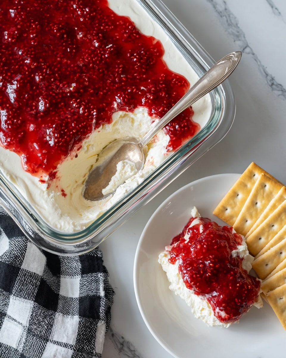 The image shows a clear glass dish with two layers: a thick, smooth white cream cheese layer at the bottom and a shiny, textured red raspberry jam layer on top, filled with raspberry seeds. A scoop is taken out of the dish, revealing the creamy layer underneath the glossy raspberry topping. Next to the dish is a white plate with a dollop of the two-layer spread mixed together, accompanied by several rectangular pale yellow crackers. The surface underneath is a white marbled texture with a black and white plaid cloth partially visible. Photo taken with an iphone --ar 4:5 --v 7