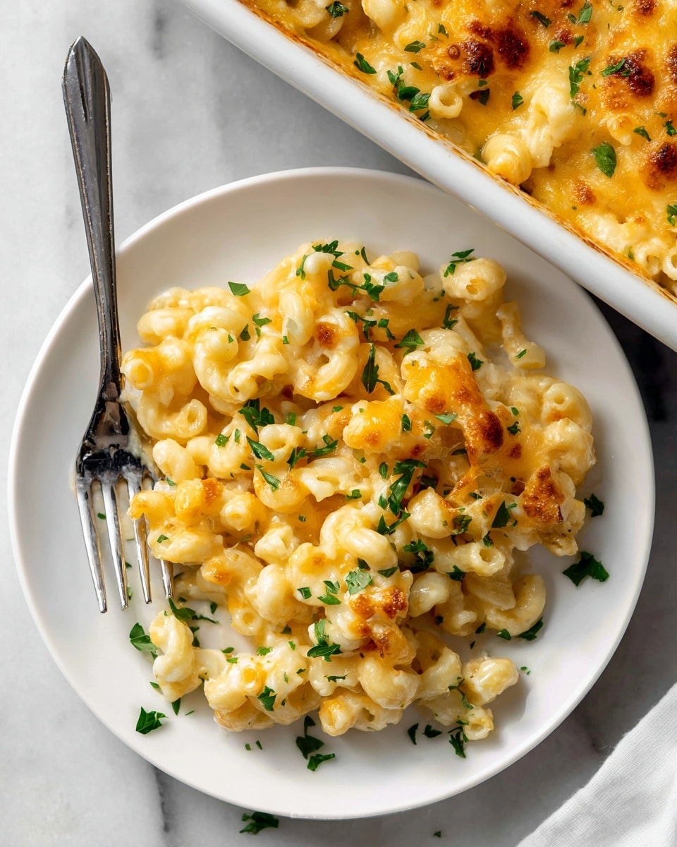 A white round plate holds a serving of macaroni and cheese with curly pasta covered in a creamy, golden-yellow cheese sauce. Some small browned spots of melted cheese are visible on top, adding texture and a slightly crispy look. Bright green parsley pieces are scattered on the pasta and around the plate for a fresh touch. A silver fork rests inside the pasta on the left side of the plate. The plate is placed on a white marbled surface, with the edge of a white rectangular baking dish filled with more macaroni and cheese visible in the top right corner. photo taken with an iphone --ar 4:5 --v 7