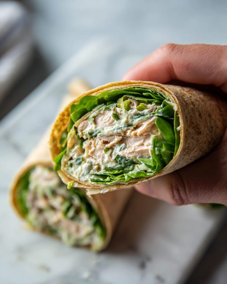 A close-up shows a hand holding a wrap with three visible layers inside: the soft light brown tortilla outside, a middle layer of creamy light green spread with small bits, and a top layer of fresh, bright green leafy lettuce. In the background, the other half of the wrap lies on a white marbled surface, showing the same filling layers. The image is focused on the wrap's cross-section, making the details of the filling clear and fresh. photo taken with an iphone --ar 4:5 --v 7