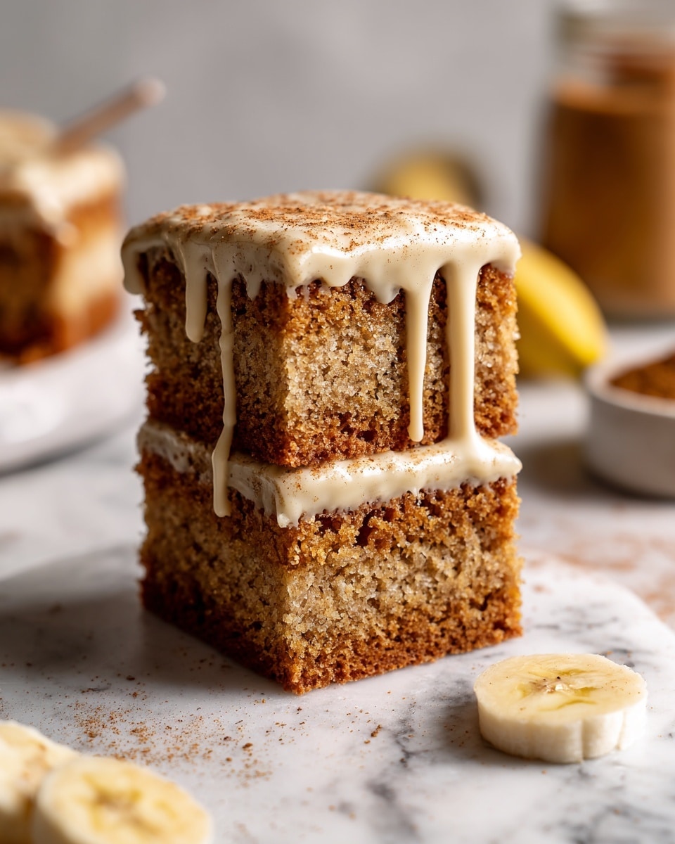 A stack of two thick, square cake pieces with a light brown, moist texture and visible crumb detail, layered with a darker cinnamon or spice mix in the middle; the top piece has a creamy, light beige icing dripping down the sides, sprinkled with fine cinnamon powder, all placed on a white marbled surface. Slices of banana lie near the cake, and blurred objects including a jar filled with brown powder appear in the background. Photo taken with an iphone --ar 4:5 --v 7
