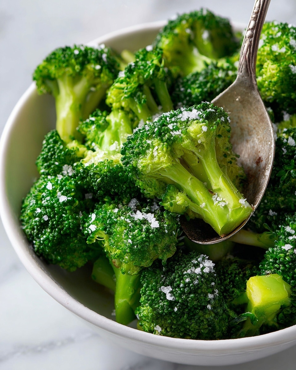 A close-up of bright green broccoli florets piled in a white bowl, each piece showing a thick stem with a dense crown of tiny buds. The broccoli looks fresh and steeped with tiny grains of coarse salt sprinkled on top, adding a slight texture contrast. A metal spoon scoops several florets from the bowl, the florets showing a vibrant mix of light green and darker shades on the buds. The scene is set on a white marbled surface, with soft light highlighting the fresh, natural texture of the broccoli. photo taken with an iphone --ar 4:5 --v 7