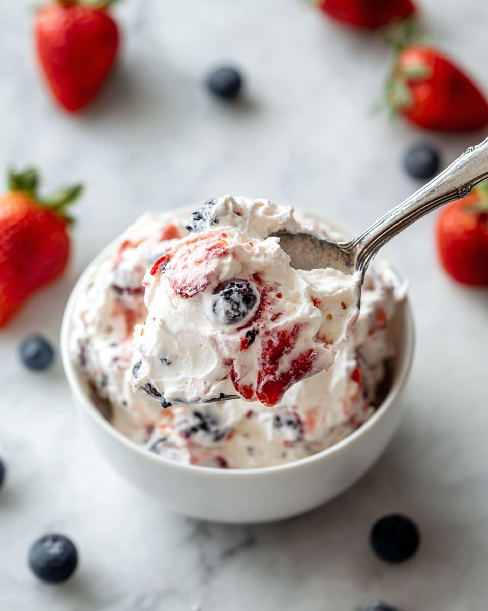 A white bowl filled with a whipped cream and fruit mixture, showing a thick layer of fluffy white cream blended with bright red strawberry pieces and dark blue blueberries evenly spread throughout. The spoon in the foreground holds a scoop of the creamy mixture with visible fruit pieces, contrasting the smooth, soft texture with the chunky fruit bits. The bowl sits on a white marbled surface with fresh strawberries and blueberries scattered nearby for added color. Photo taken with an iphone --ar 4:5 --v 7
