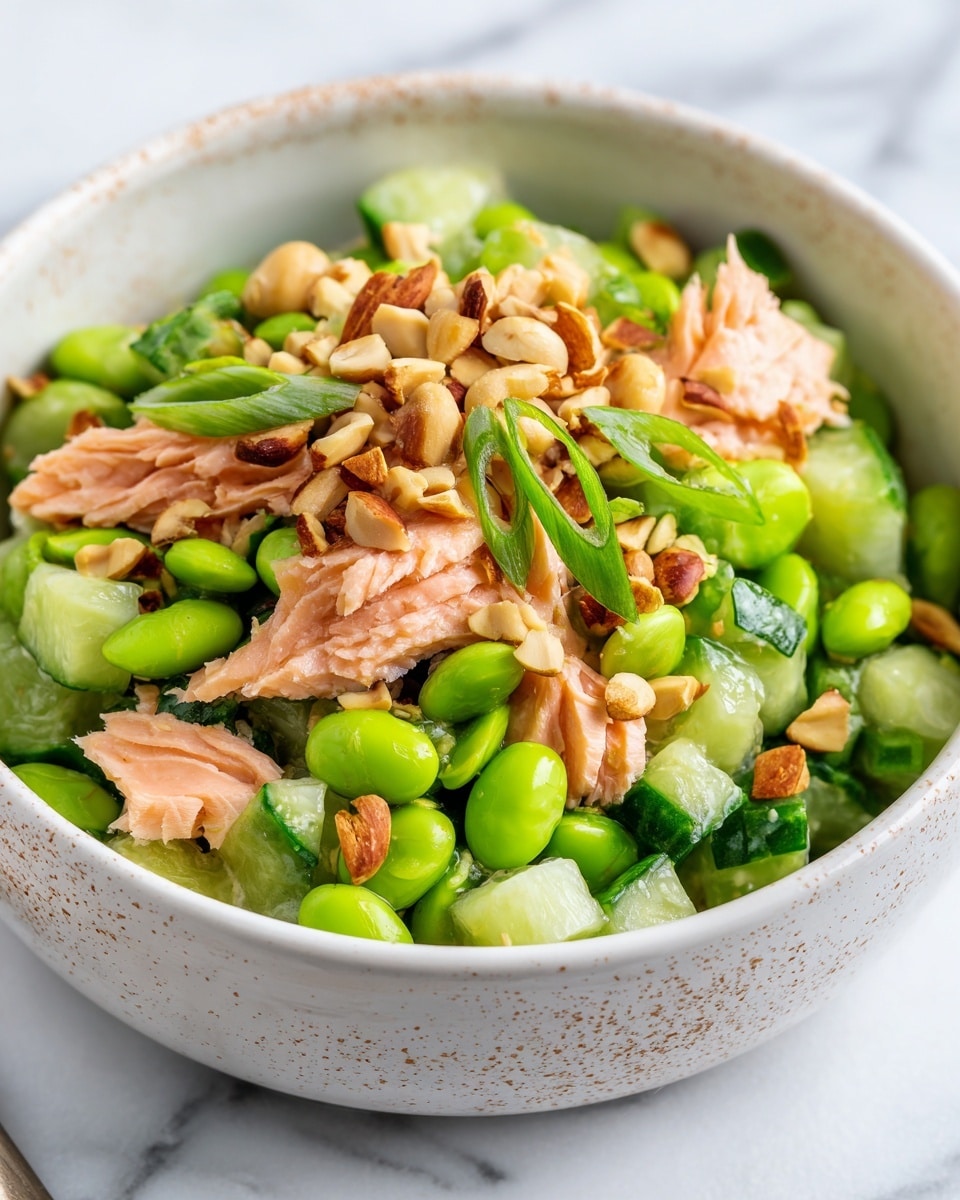 The image shows a bowl filled with a fresh, mixed salad. The salad has several layers: at the bottom are chunks of light green cucumber pieces with a slightly watery texture, mixed with bright green edamame beans that have a smooth texture. Scattered throughout are small pieces of light pink cooked salmon with a soft, flaky look. On top, a sprinkle of roughly chopped light brown nuts adds a crunchy texture, and thin slices of bright green spring onions are dotted across for a fresh look. The bowl is white with a light brown speckled pattern on the inside, sitting on a white marbled surface. Photo taken with an iphone --ar 4:5 --v 7