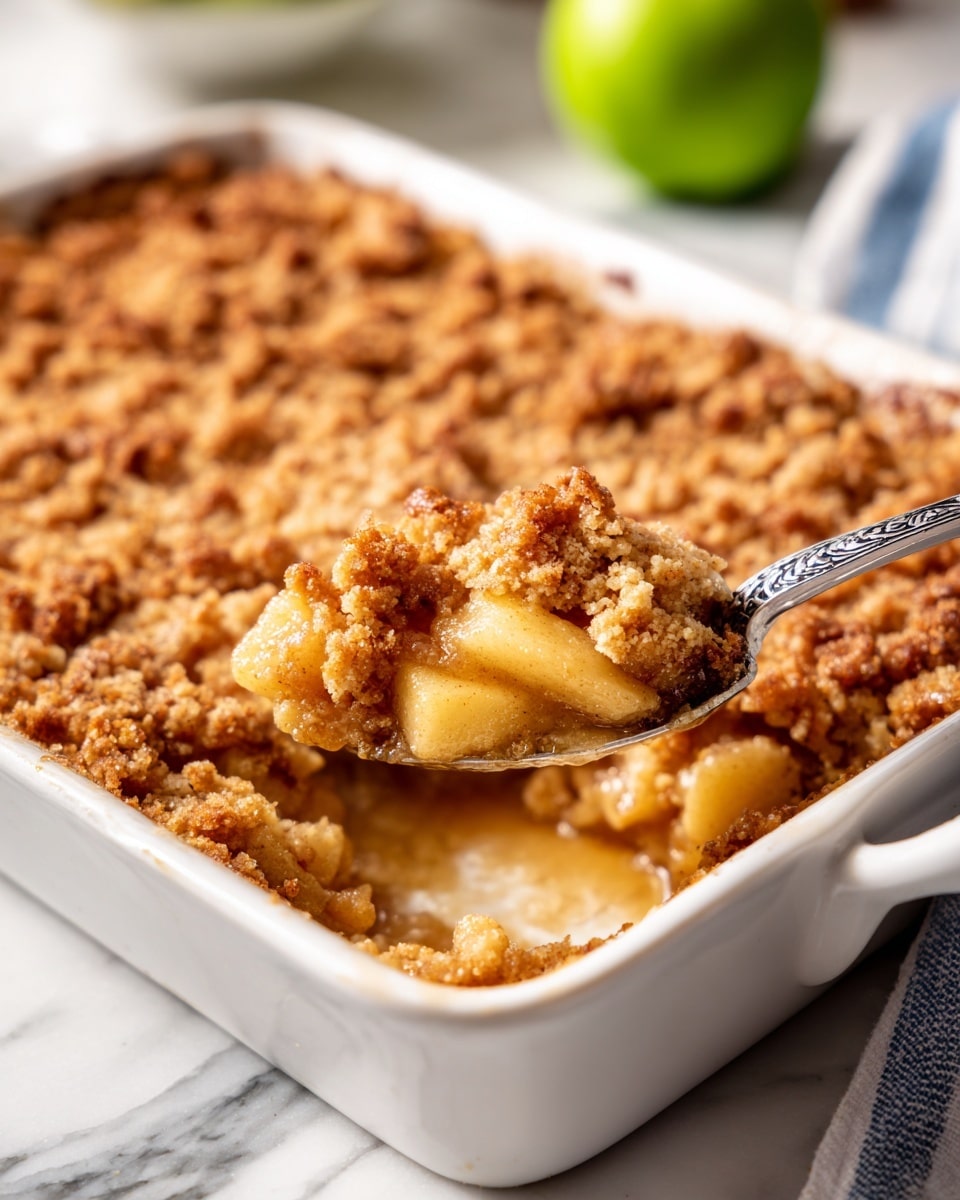 A close-up view of a white rectangular baking dish filled with a golden brown apple crumble. The crumble has a crumbly, textured top layer with uneven small chunks and a warm toasted color. Below this crumbly layer, a glossy, thick apple filling is partially visible where a spoon scoops it out. The spoon has a detailed, ornate silver handle and sits in the middle of the dish. The baking dish is on a white marbled surface with a blurred green apple and a blue and white striped cloth nearby. Photo taken with an iphone --ar 4:5 --v 7