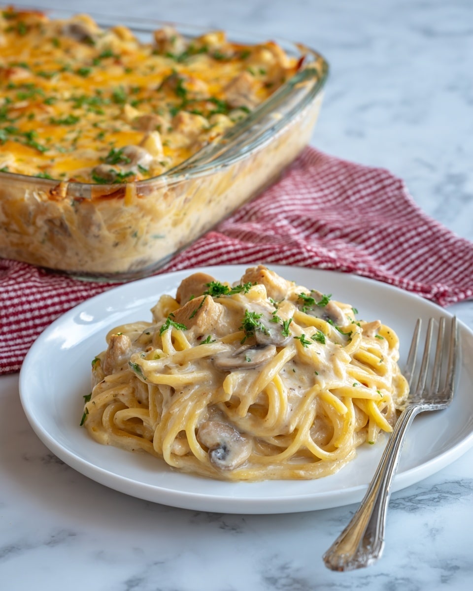 A white plate holds a large serving of creamy pasta with visible pieces of chicken mixed in. The pasta is covered in a thick, smooth, light beige sauce, garnished with small green parsley flakes. Behind the plate is a clear glass baking dish filled with the same pasta dish, topped with a golden brown layer of melted cheese. The baking dish sits on a soft red and white checked cloth on a white marble surface. A silver fork rests next to the plate on the right. photo taken with an iphone --ar 4:5 --v 7