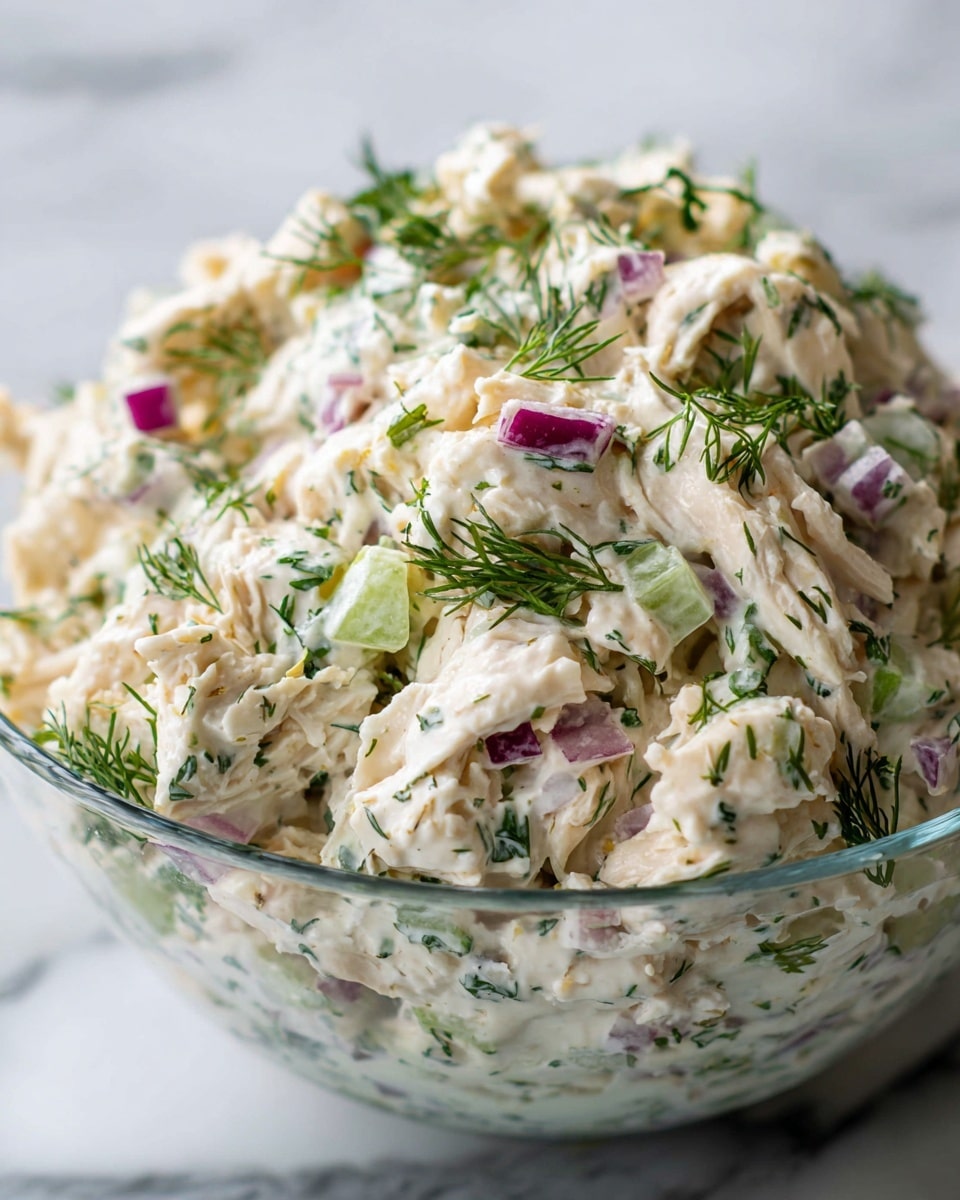 This close-up image shows a creamy salad in a clear bowl on a white marbled surface. The salad has three main layers: a base of pale shredded chicken mixed smoothly with a creamy white dressing, scattered pieces of chopped celery adding light green color and crunchy texture, and small chunks of purple-red onion mixed throughout. The salad is sprinkled with delicate green dill leaves that float on top, giving a fresh and colorful look. The textures vary from soft creamy chicken to crisp celery, all coated evenly in dressing. Photo taken with an iphone --ar 4:5 --v 7