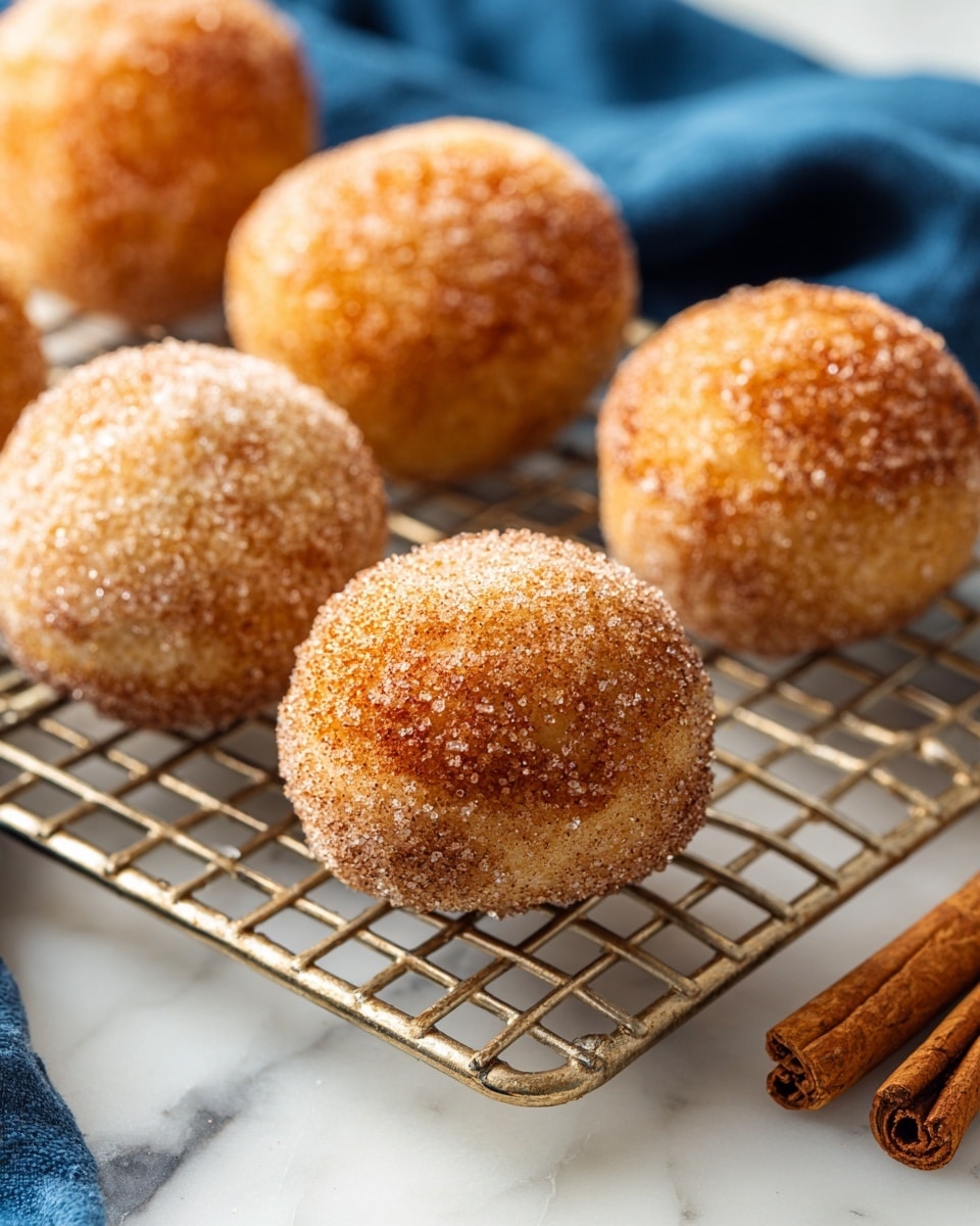 The image shows small round doughnuts arranged on a silver wire cooling rack. Each doughnut is covered with a layer of sugar, giving them a grainy texture and a light brown color. The surface of the doughnuts appears soft and fluffy. The rack sits on a surface with a white marbled texture, and there is a small piece of cinnamon stick nearby. In the corner of the image, part of a blue cloth is visible, adding a touch of color to the scene. photo taken with an iphone --ar 4:5 --v 7