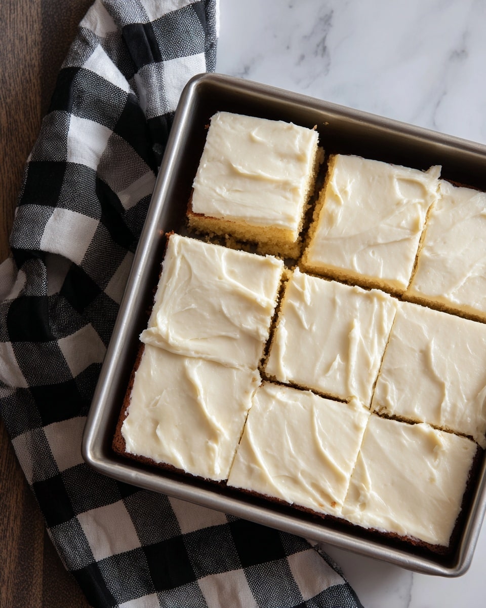 A metal baking pan holds twelve square cake pieces arranged in three rows of four, each topped with a thick layer of smooth, white frosting with slight texture marks. The frosting covers the top fully, and a few edges show a hint of soft yellow cake beneath. One piece is missing from the top left corner, revealing a clean cut edge. The pan rests on a black and white checkered cloth placed over a wood-textured surface, all against a white marbled texture background. Photo taken with an iphone --ar 4:5 --v 7