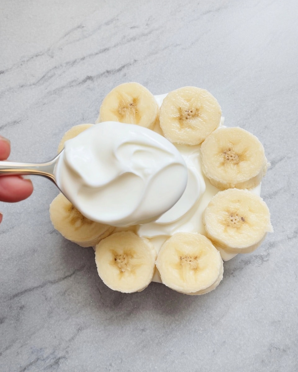 The image shows round banana slices arranged closely in a small circle on a white marbled surface, forming the base layer. On top of the banana slices, a spoon held by a woman's hand is spreading a thick, smooth layer of white yogurt, covering the center of the bananas while some slices remain visible at the edges. The yogurt layer has a creamy texture and bright white color that contrasts with the pale yellow bananas. Photo taken with an iphone --ar 4:5 --v 7
