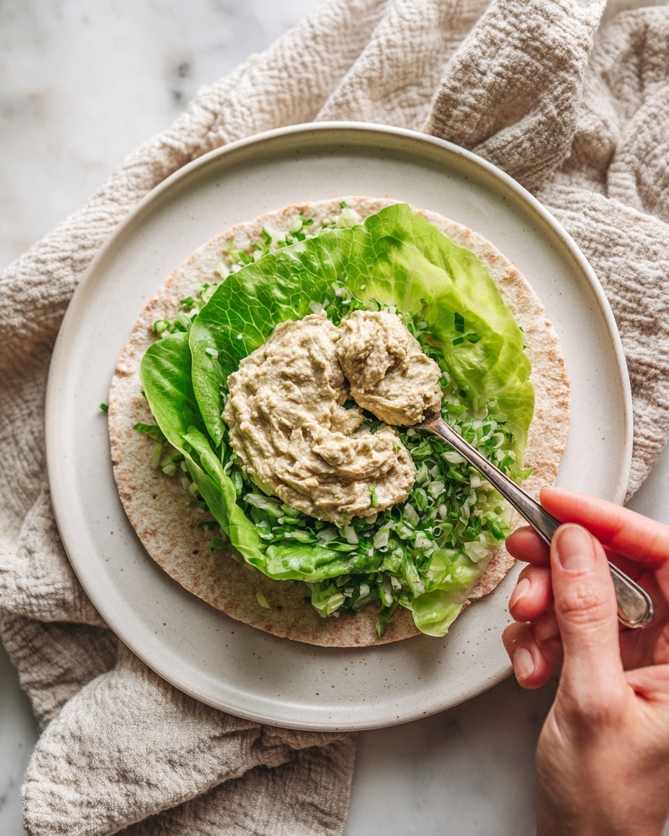 A flat light brown bread with an uneven surface is laid out on a white plate on a white marbled surface. On top of the bread, there is a layer of finely chopped fresh green lettuce spread in the center. Above the lettuce, a woman’s hand holds a spoon with a scoop of thick, pale green mixture being placed in the middle of the lettuce. A textured cloth in light color is seen blurred in the background. Photo taken with an iphone --ar 4:5 --v 7