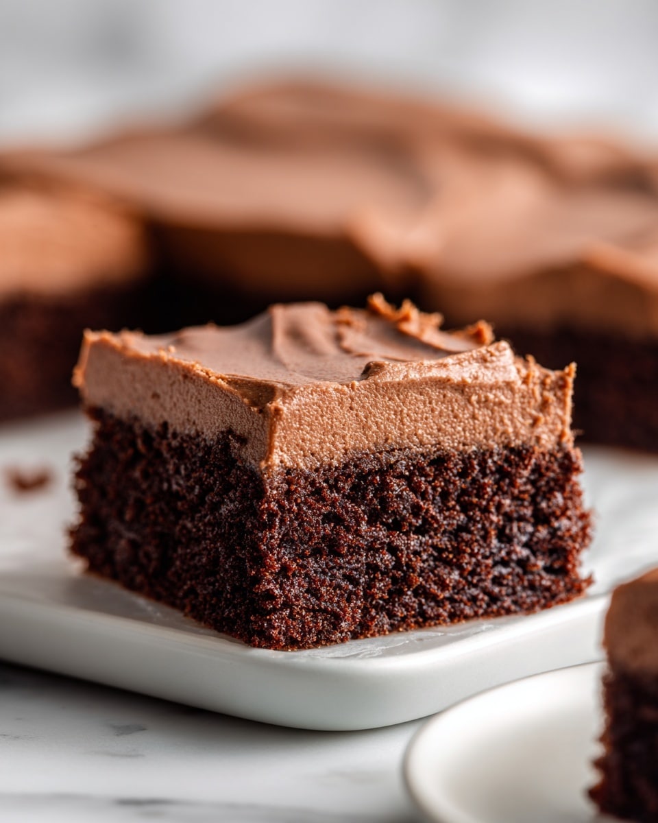 A close-up of a square chocolate cake piece held above a white baking tray with the rest of the cake inside, showing one thick bottom layer of dark brown chocolate cake and one smooth top layer of lighter brown chocolate frosting. The cake is partly cut into squares, and a white plate with another cake piece sits blurred in the background on a white marbled surface. The texture of the cake layer looks soft and slightly crumbly, while the frosting looks creamy and spread evenly with small rough edges on top. photo taken with an iphone --ar 4:5 --v 7