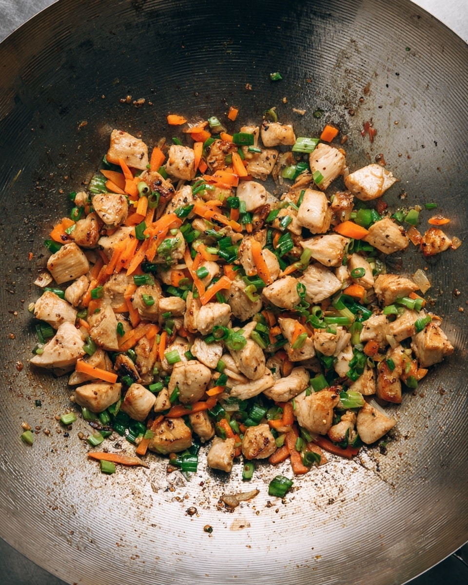 Inside a large metal wok, there is a mix of small light brown cubes of cooked chicken, thin orange strips of carrots, and small green pieces of chopped spring onions, all evenly spread and slightly browned from cooking. The wok’s surface shows some cooked oil and darker sear spots, adding texture and contrast to the mix of ingredients. The light and dark colors blend naturally with the shiny metal base. Photo taken with an iphone --ar 4:5 --v 7