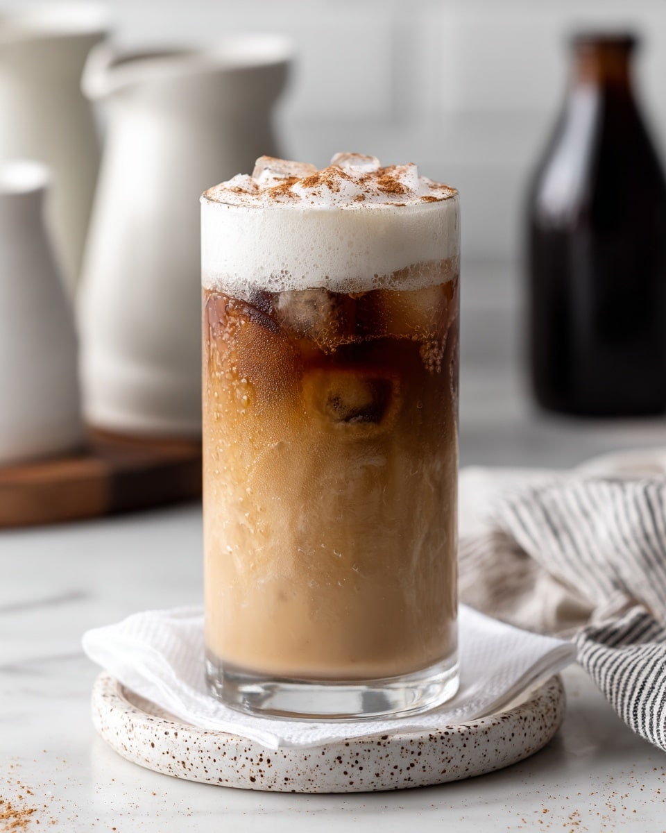 A tall clear glass holds an iced coffee drink with three main layers: the bottom layer is a light tan coffee with ice cubes visible inside, the middle layer is a slightly darker coffee, and the top layer has thick white frothy foam sprinkled with brown cinnamon powder. The glass is placed on a white speckled circular coaster with a white napkin underneath, all set on a white marbled surface. In the background, there are blurred white ceramic containers and a dark bottle, with a striped cloth on the right side. photo taken with an iphone --ar 4:5 --v 7