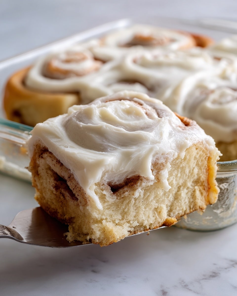 The image shows a square slice of cinnamon roll with two visible layers: the bottom layer is a soft, light beige dough with swirls of light brown cinnamon filling, and the top layer is thick, smooth, creamy white frosting evenly spread with visible soft peaks. The slice is held by a spatula in a clear baking dish placed on a white marbled surface. Photo taken with an iphone --ar 4:5 --v 7