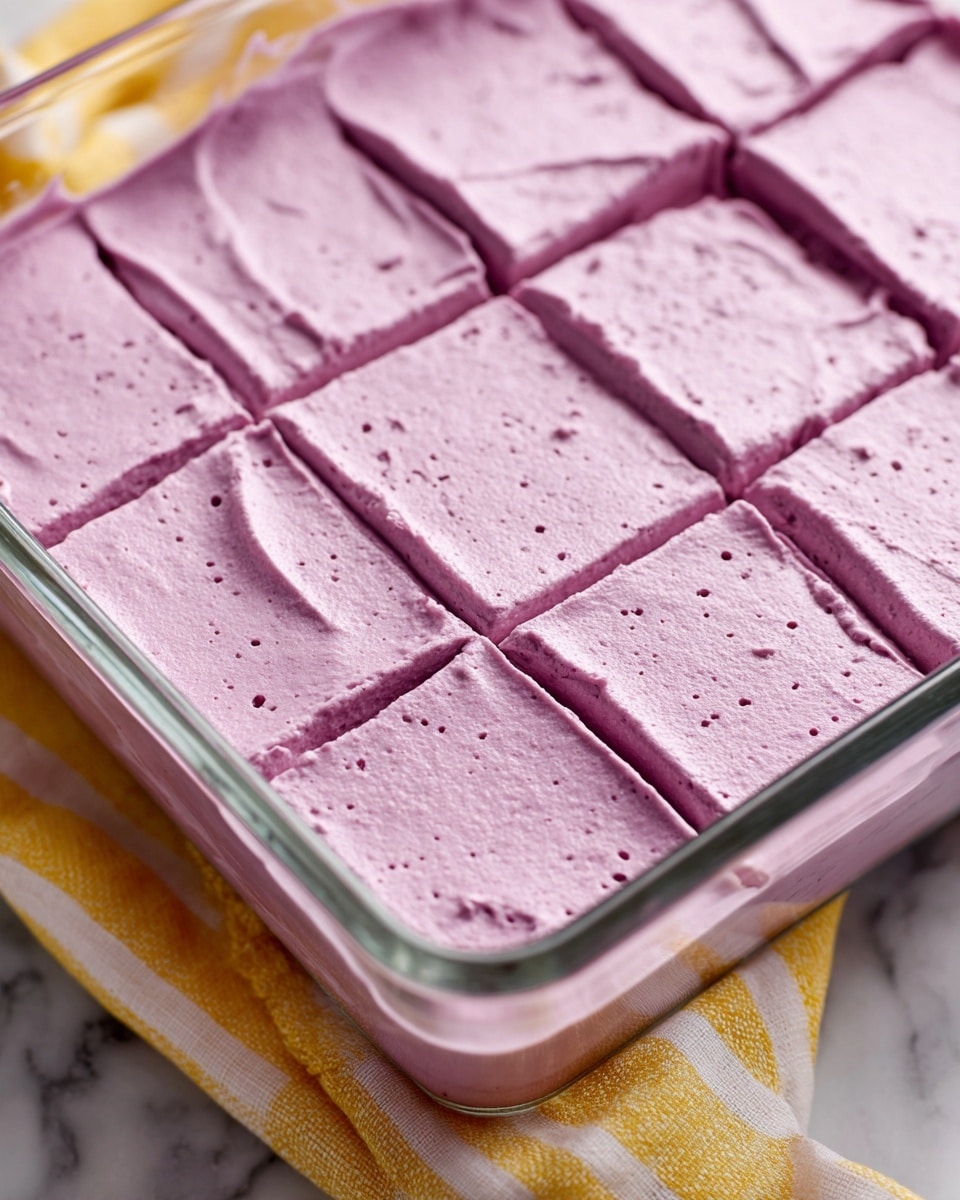 A close-up view of a glass baking dish filled with a thick, smooth layer of purple cream. The surface is slightly textured with small specks of darker purple and lightly scored into squares, creating a grid pattern. The dish sits on a white marbled textured surface with a yellow and white striped cloth partially visible underneath. The purple cream looks rich and evenly spread with soft peaks and slight folds. Photo taken with an iphone --ar 4:5 --v 7