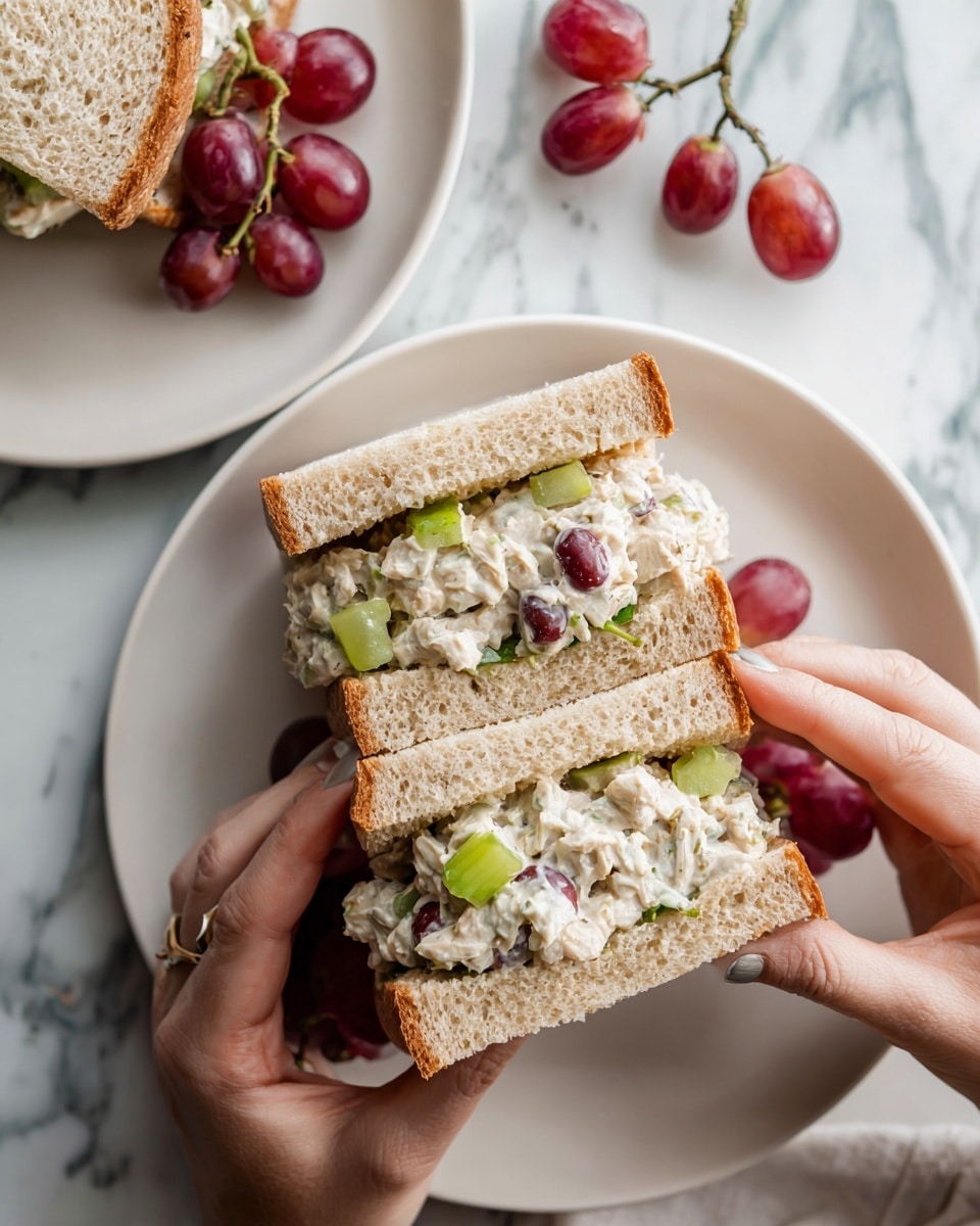 A sandwich held by two hands shows three layers: the top and bottom layers are slices of light brown bread with a soft texture, and the middle layer is a mix of creamy chicken salad with white and light yellow tones, pieces of green celery, and red grape halves. The sandwich rests over a white plate set on a white marbled surface with a few loose red grapes scattered nearby. In the background, a similar sandwich and grapes sit on another white plate. Photo taken with an iphone --ar 4:5 --v 7
