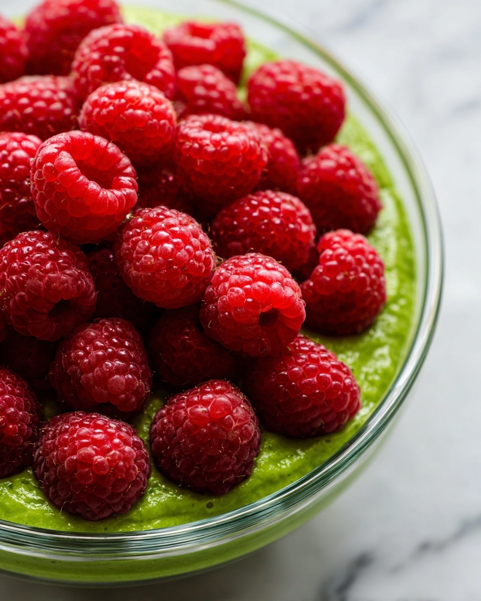 A close-up view of a clear glass bowl filled with a bright green thick paste with a creamy texture at the bottom and sides, topped with a layer of fresh, plump, and shiny red raspberries covering the green layer almost completely. The raspberries have a natural bumpy surface and rich color, arranged in a slightly uneven cluster. The bowl is set on a white marbled surface with subtle veins for contrast. Photo taken with an iphone --ar 4:5 --v 7