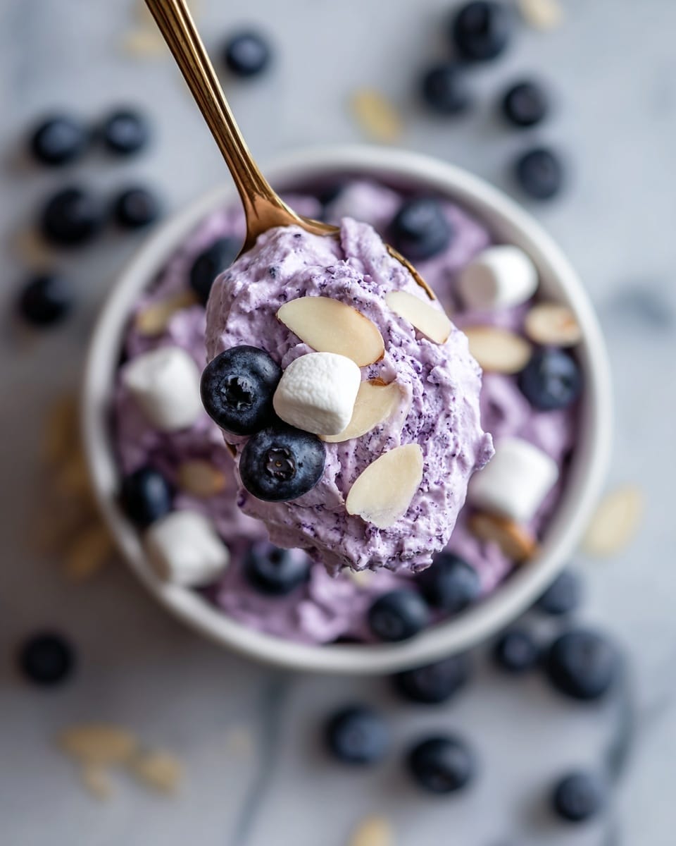 A close-up of a spoon holding a serving of light purple whipped dessert mixed with whole blueberries and topped with thin almond slices and one small white marshmallow. In the background, the dessert fills a white bowl and has more blueberries and marshmallows scattered on top. The surface is a white marbled texture and the spoon has a golden handle. Photo taken with an iphone --ar 4:5 --v 7