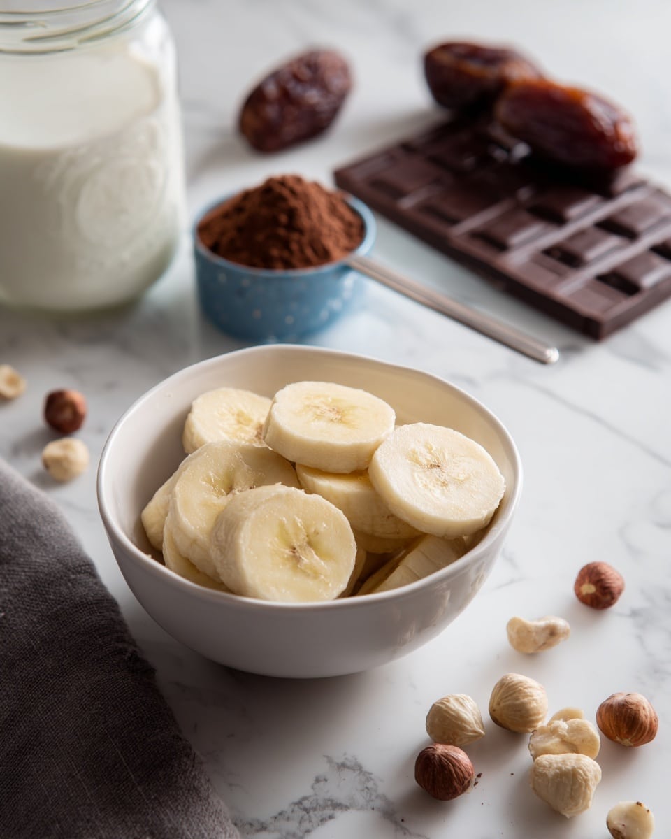 A white small bowl filled with round slices of pale yellow banana is in the front. Behind the bowl on a white marbled surface, there is a clear jar filled with white milk on the left side, a blue measuring spoon holding brown cocoa powder in the middle, two brown dates further back, and a bar of dark chocolate on the right. Scattered on the surface near the bowl are light brown hazelnuts. The photo taken with an iphone --ar 4:5 --v 7