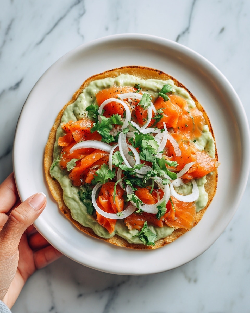 A white plate on a white marbled surface holds a single crispy golden brown tortilla as the base. On top of this layer is a spread of light green guacamole, smooth in texture, followed by bright orange slices of cooked salmon arranged evenly. Over the salmon, thin, curly white onion slices are scattered along with small fresh green cilantro leaves. A woman's hand is gently holding the edge of the plate. photo taken with an iphone --ar 4:5 --v 7
