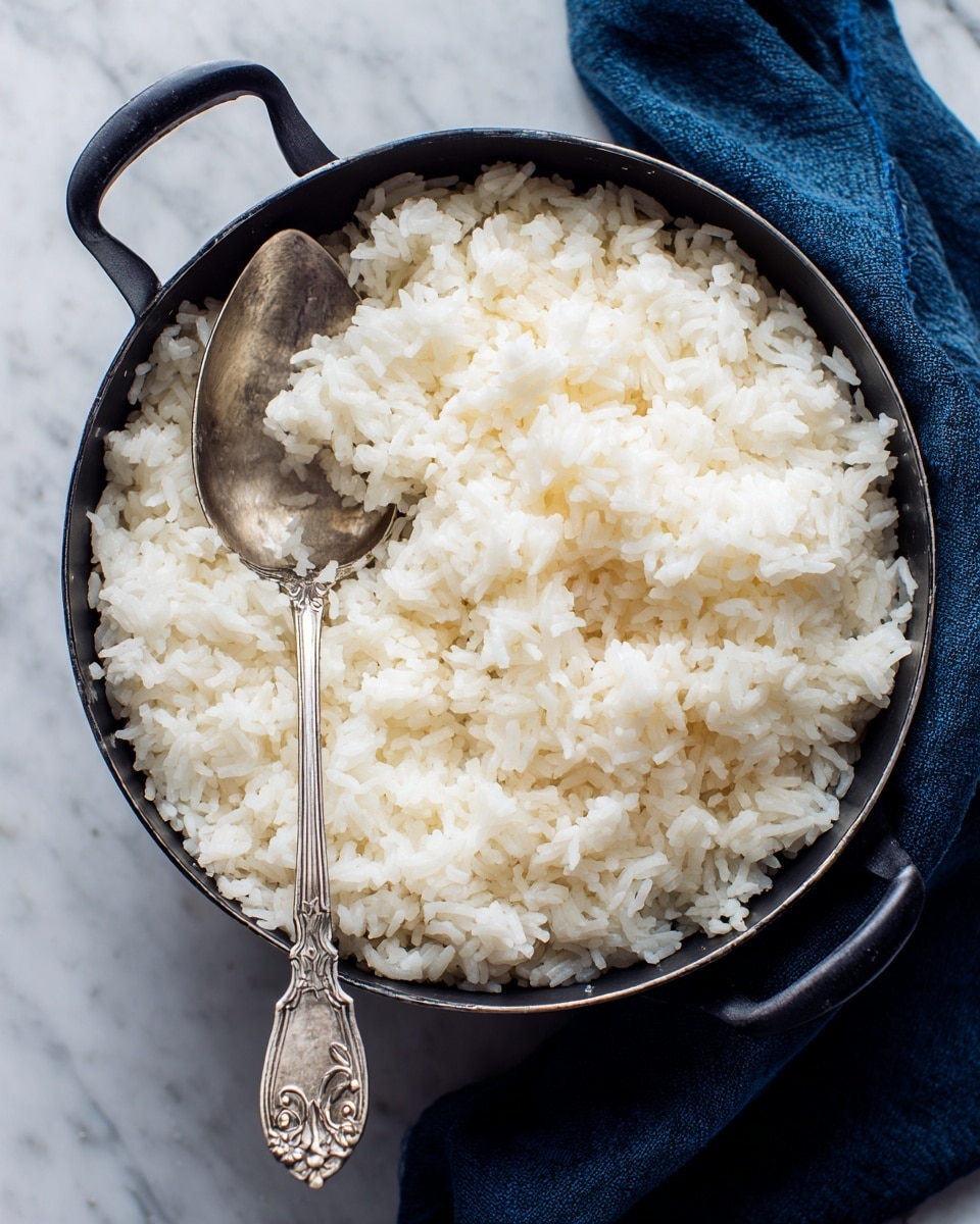 A close-up of a black pot filled with cooked white rice, the rice grains appear soft and slightly fluffy, covering the entire visible area inside the pot. A silver spoon with an ornate handle is partially submerged in the rice on the left side, resting against the pot's edge. The pot is placed on a white marbled surface, with a dark blue cloth partially visible underneath on the right side. photo taken with an iphone --ar 4:5 --v 7
