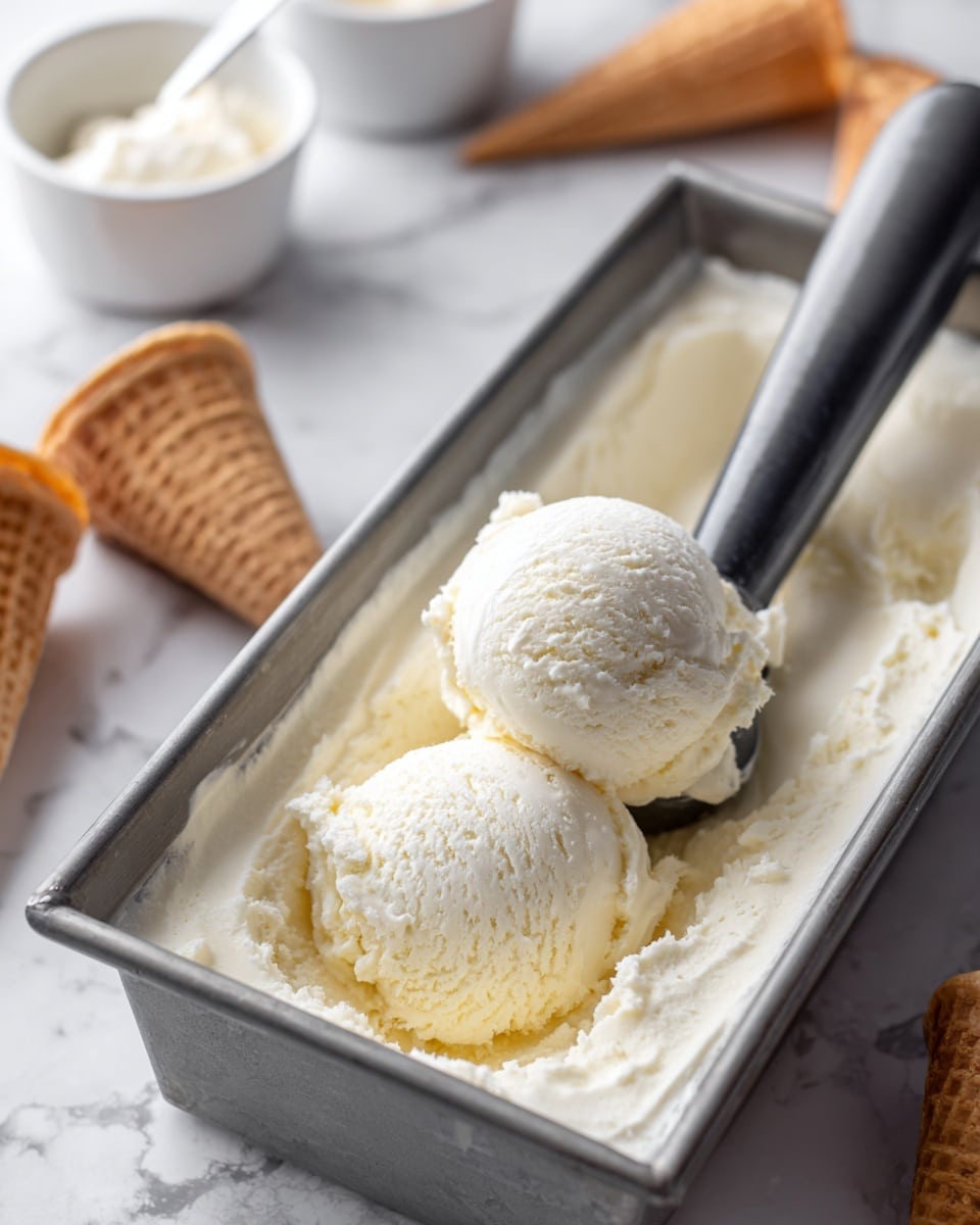 A close-up view of a metal rectangular container filled with creamy, off-white ice cream. There are two scoops of ice cream in the container, with one scoop held by a dark gray ice cream scooper positioned diagonally across the container. The ice cream has a smooth but slightly textured surface. In the soft-focused background, there are some white bowls with spoons and several waffle cones standing upright on a white marbled surface. photo taken with an iphone --ar 4:5 --v 7