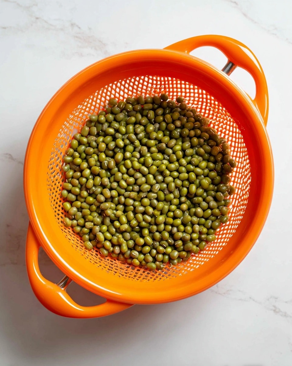 A close-up view of a bright orange plastic strainer filled with small green mung beans. The strainer has a grid pattern and two rounded handles, one on each side. The mung beans inside show a variety of green shades, from light to dark, with a smooth texture. The strainer sits on a white marbled surface, adding a clean and bright background to the image. Photo taken with an iphone --ar 4:5 --v 7
