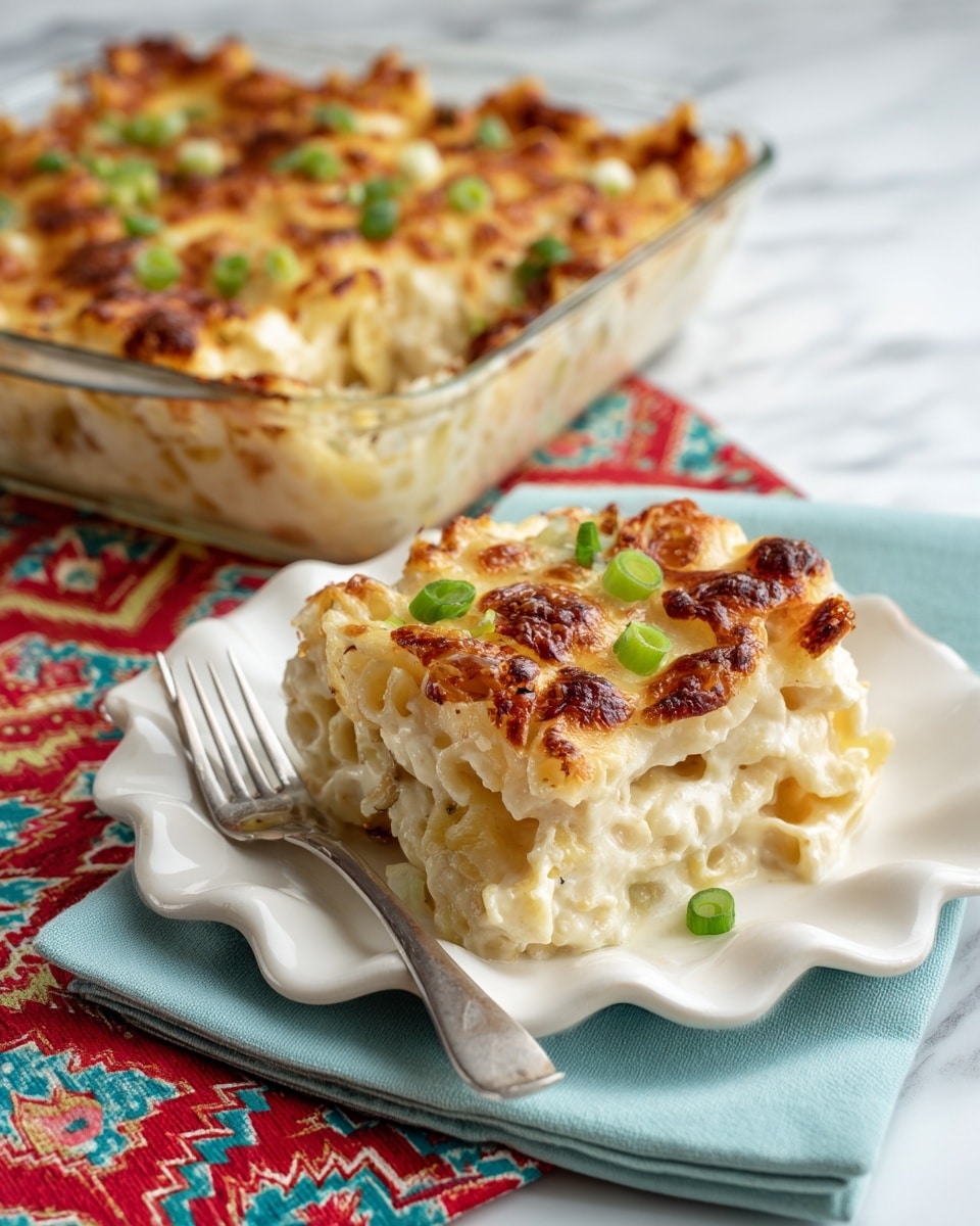 A piece of baked pasta casserole served on a white scalloped plate, showing several layers including a golden brown melted cheese top with small green onion slices and a few darker toasted spots, the pasta underneath looks creamy and slightly textured with sauce. The casserole dish in the background is clear glass, filled with a thick layer of pasta and cheese, also topped with green onions, the edges are slightly browned. The plate sits on a light blue cloth napkin with a silver fork next to it, placed on a white marbled surface with a red and multicolor zigzag patterned fabric underneath. Photo taken with an iphone --ar 4:5 --v 7