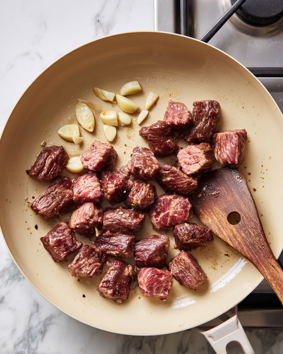 Inside a beige pan, small cubes of browned meat with pink centers are gathered mostly on the left side, showing a juicy texture. Near the cubes, a few light yellow garlic cloves are scattered with slight browning on their edges. On the right side of the pan, a wooden spatula rests, showing a dark wood color with a small hole near its edge. The pan sits on a stovetop with visible burners below, and the background is a white marbled texture. photo taken with an iphone --ar 4:5 --v 7