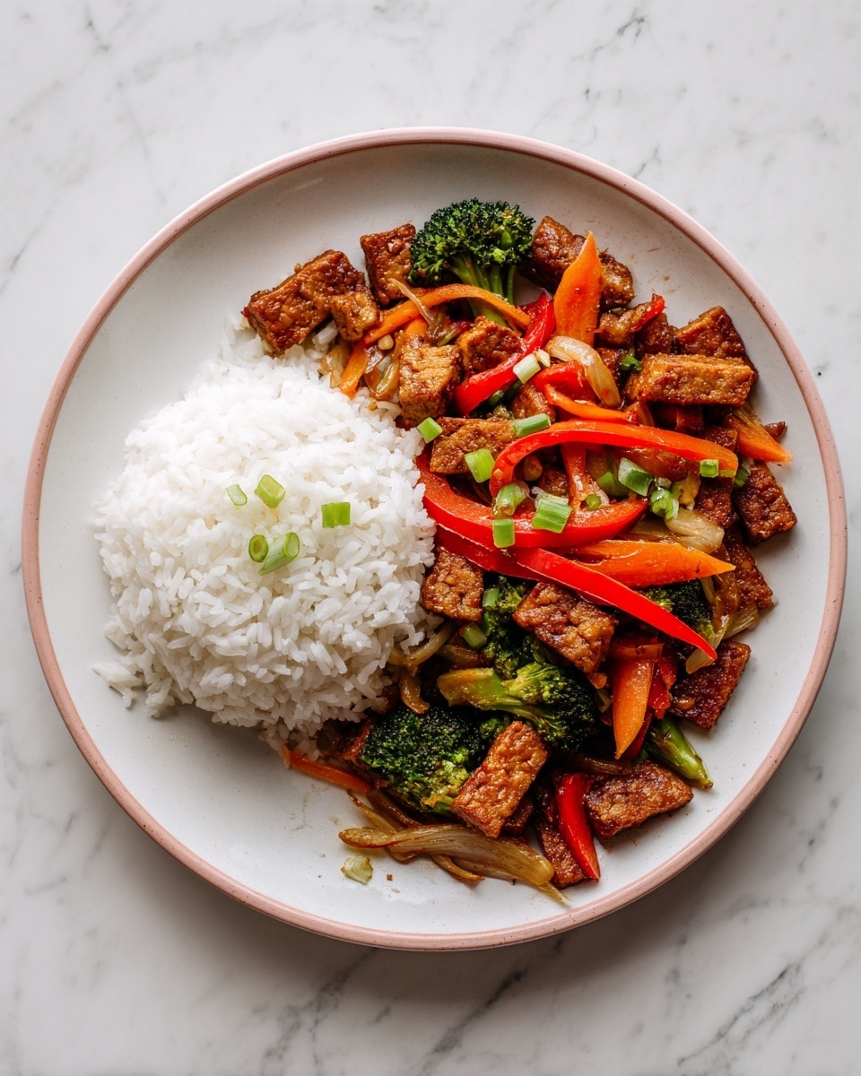 The image shows a white plate with a peach rim, placed on a white marbled surface. On the left side of the plate, there is a mound of plain white rice with a soft texture. On the right side, there is a colorful stir-fried mixture consisting of irregular squares of brown tempeh, thin slices of bright red bell peppers, small green broccoli florets, thin orange carrot sticks, and pieces of translucent cooked onion. The dish is garnished with small pieces of green onion scattered on top. The food is neatly placed with the rice and vegetable-tempeh mix side by side, showing a contrast of white and warm colors. photo taken with an iphone --ar 4:5 --v 7